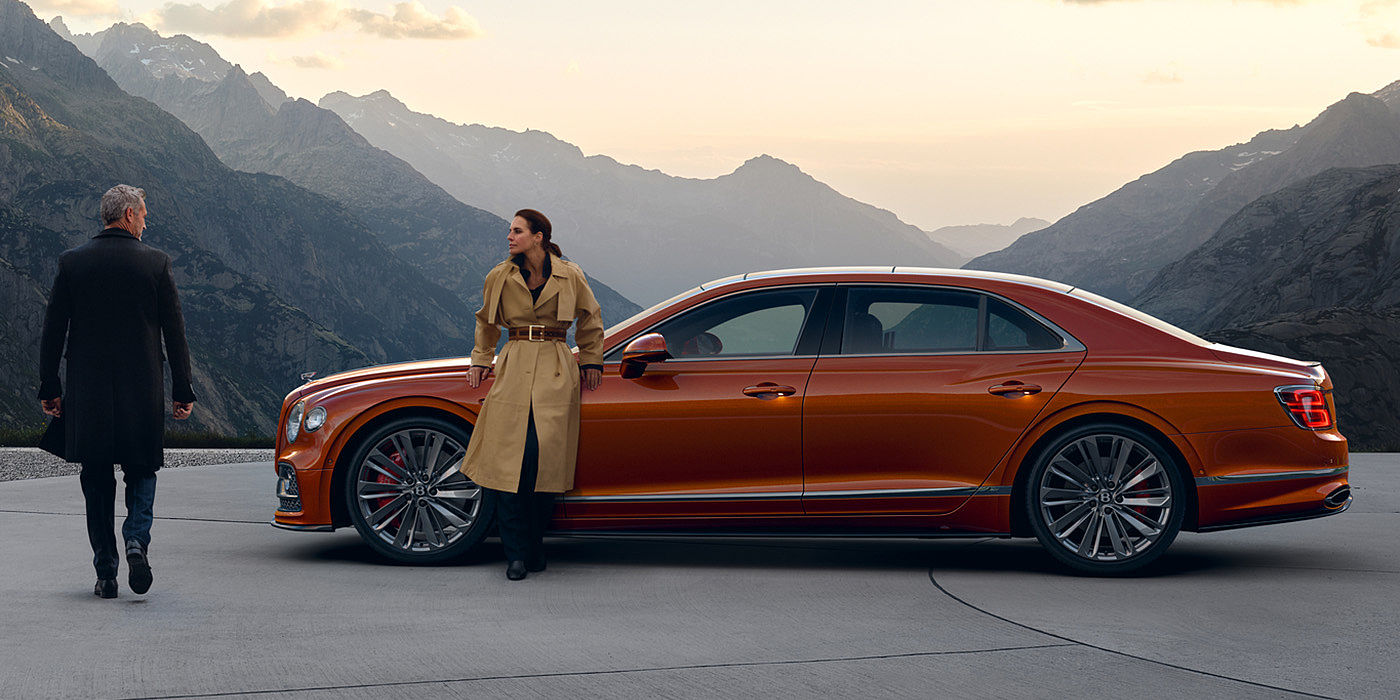 Bentley Zurich Bentley Flying Spur Speed parked in Orange Flame coloured exterior parked, with mountainous background and two people in view.