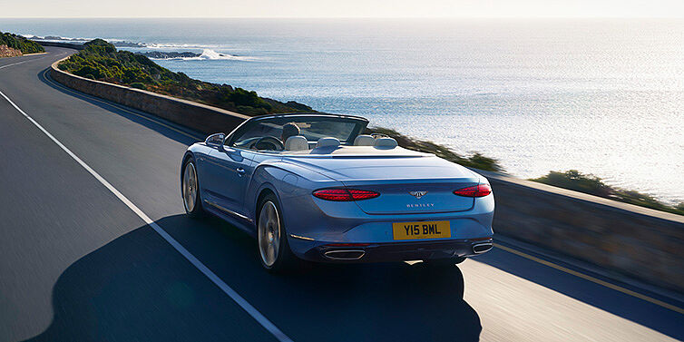 Bentley Zurich New Bentley Continental GTC Azure convertible rear view in Light Windsor blue paint with 22 inch ten spoke wheels driving dynamically on an ocean road in the sunshine