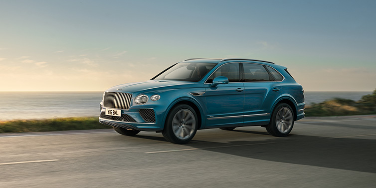 Bentley Zurich Bentley Bentayga Azure SUV in Topaz blue paint driving dynamically by the ocean with 22 inch 10 spoke directional wheels