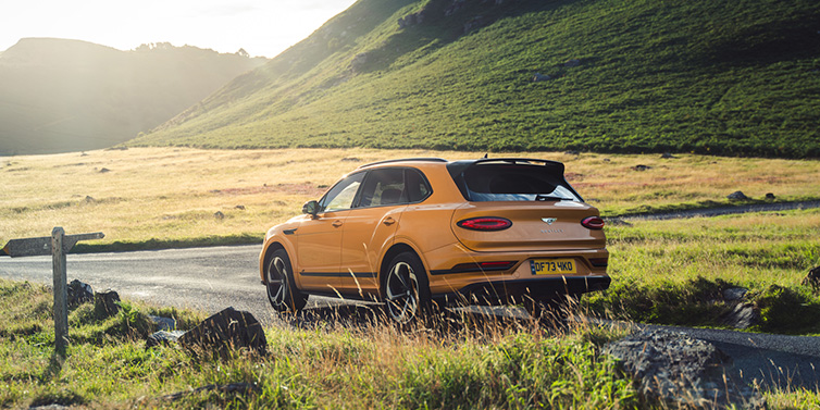 Bentley Zurich Bentley Bentayga S SUV rear three quarter in Sunburst Gold paint on a road surrounded by green hills