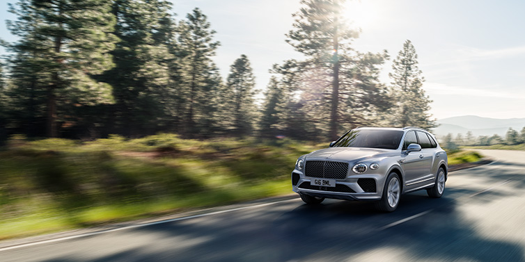 Bentley Zurich Bentley Bentayga SUV front three quarter in Moonbeam silver paint driving dynamically along a tree-lined road