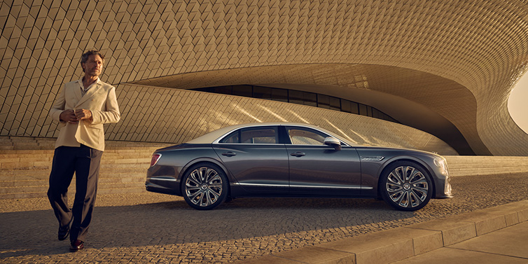 Bentley Zurich Bentley Flying Spur Mulliner sedan in Rose Gold over Magnetic duotone paint parked by a curved wall with person in suit jacket standing on the left